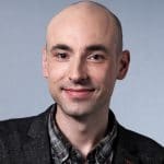 Porträt eines Mannes mit Glatze, lächelnd, in einem grauen Blazer und karierten Hemd, vor einem neutralen Hintergrund.