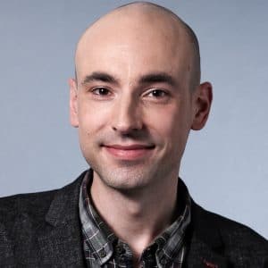 Porträt eines Mannes mit Glatze, lächelnd, in einem grauen Blazer und karierten Hemd, vor einem neutralen Hintergrund.