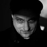 Bernd Arnold Porträt eines Mannes mit einem ernsten Blick, teilweise im Schatten, trägt eine schwarze Jacke und eine Mütze.