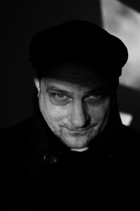 Bernd Arnold Porträt eines Mannes mit einem ernsten Blick, teilweise im Schatten, trägt eine schwarze Jacke und eine Mütze.