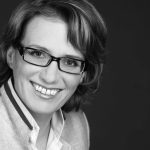 Kerstin Plehwe Porträt einer lächelnden Frau mit Brille, kurzen Haaren und einem Blazer, vor einem dunklen Hintergrund.