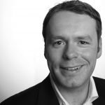 Maik Bohne Schwarz-weiß Porträt eines lächelnden Mannes in Anzug vor hellem Hintergrund.