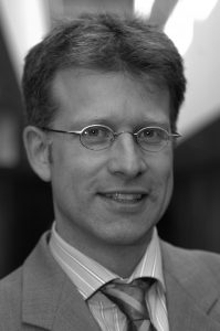 Carel Mohn Porträt eines Mannes mit Brille, lächelnd, in einem Anzug mit gestreifter Krawatte, vor einem unscharfen Hintergrund.