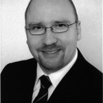 Schwarz-weiß Porträt eines Mannes mit Brille, Glatze und Anzug, lächelnd in die Kamera.