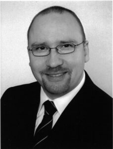 Schwarz-weiß Porträt eines Mannes mit Brille, Glatze und Anzug, lächelnd in die Kamera.