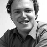 Mario Voigt Porträt eines lächelnden Mannes mit lockigem Haar, der ein Hemd trägt und an einem Tisch sitzt. Schwarz-weiß-Foto.