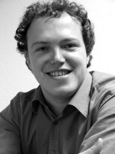 Mario Voigt Porträt eines lächelnden Mannes mit lockigem Haar, der ein Hemd trägt und an einem Tisch sitzt. Schwarz-weiß-Foto.