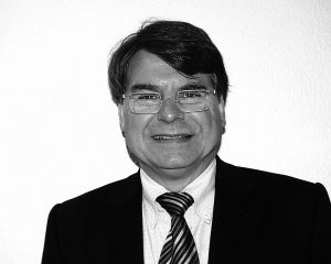 Porträt eines Mannes mit Brille, dunklem Anzug und gestreifter Krawatte vor einer hellen Wand.