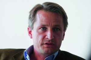 Porträt eines Mannes mit kurzen, blonden Haaren und blauen Augen, der ernst in die Kamera schaut.