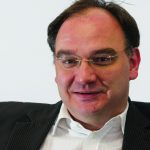 Nico Fried Porträt eines Mannes mit Brille, in Anzug und Hemd, lächelnd vor einem hellen Hintergrund.