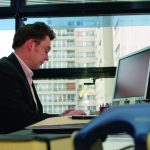 Thymian Bussemer Mann in Anzug sitzt an Schreibtisch, tippt auf Computer, Blick aus Fenster auf moderne Gebäude.