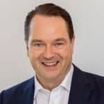 Lächelnder Mann in Anzug vor neutralem Hintergrund, mit kurzen, dunklen Haaren.