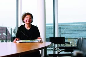 Porträt von Brigitte Fehrle, sitzend an einem Tisch mit Zeitungen, in einem modernen Raum mit großen Fenstern.