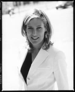 Porträt einer lächelnden Frau mit blonden Haaren, die einen weißen Blazer trägt, vor einem hellen Hintergrund.