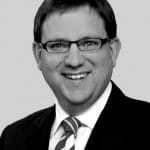 Porträt eines lächelnden Mannes mit Brille, Anzug und gestreifter Krawatte vor grauem Hintergrund.