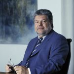 Kurt Beck Portrait von Kurt Beck in Anzug, sitzend an einem Tisch, mit Brille in der Hand, vor abstraktem Hintergrund.