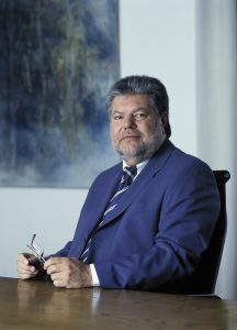 Kurt Beck Portrait von Kurt Beck in Anzug, sitzend an einem Tisch, mit Brille in der Hand, vor abstraktem Hintergrund.
