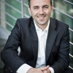 Thomas Jarzombek Mann im Anzug sitzt lächelnd auf einer Treppe, mit verschränkten Händen und modernem Hintergrund.