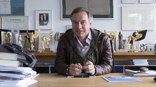 Mann sitzt an einem Schreibtisch, umgeben von Auszeichnungen und Dokumenten, mit einem nachdenklichen Ausdruck.