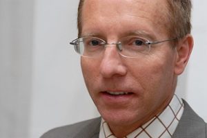 Porträt eines Mannes mit Brille, blonden Haaren, in einem grauen Anzug und karierten Hemd, vor einem hellen Hintergrund.