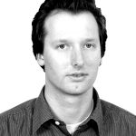 Jörg-Uwe Nieland Schwarz-weiß-Porträt eines Mannes mit dunklen Haaren, der in einem karierten Hemd in die Kamera schaut.