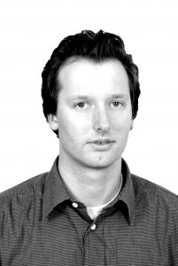Jörg-Uwe Nieland Schwarz-weiß-Porträt eines Mannes mit dunklen Haaren, der in einem karierten Hemd in die Kamera schaut.