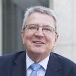 Achim Moeller Porträt eines lächelnden Mannes mit grauen Haaren, Brille und Anzug, vor einem hellen Hintergrund.