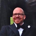 Christoph Tophinke Mann mit Brille und Glatze, trägt einen Anzug mit Fliege, lächelt vor einem dunklen Hintergrund.