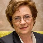 Christine Bauer-Jelinek Porträt einer Frau mit braunen, lockigen Haaren, Brille und einem freundlichen Lächeln, in formeller Kleidung.