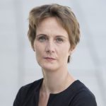 Melanie Ahlemeier Porträt einer Frau mit kurzen, braunen Haaren, in schwarzem Oberteil, mit ernstem Gesichtsausdruck vor einem hellen Hinte...