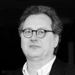 Lutz Meyer Porträt eines Mannes mit Brille und lockigem Haar, der in die Kamera schaut, vor einem dunklen Hintergrund.
