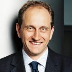 Alexander Graf Lambsdorff Porträt eines lächelnden Mannes mit kurzen, lockigen Haaren, in einem Anzug mit Krawatte, vor neutralem Hintergrund.
