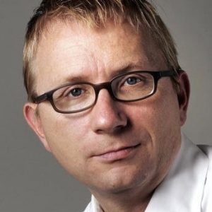 Porträt eines Mannes mit blonden Haaren und Brille, der direkt in die Kamera schaut.