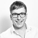 Alexander Hauk Schwarz-weiß Porträt eines lächelnden Mannes mit Brille und kurzen Haaren, trägt ein helles Hemd.