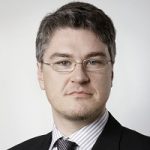 Porträt eines Mannes mit Brille, kurzen grauen Haaren, Anzug und gestreifter Krawatte vor neutralem Hintergrund.