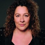 Ulrike Simon Porträt einer Frau mit lockigem, braunem Haar, die direkt in die Kamera schaut, vor dunklem Hintergrund.