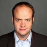 Robin Alexander, Porträt eines Mannes mit kurzen Haaren, neutralem Gesichtsausdruck, in einem dunklen Anzug und hellblauer Hemd.