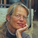 Inse Geismar Porträt einer Frau mit Brille, die lächelt und ihren Kopf auf die Hand stützt, im Hintergrund unscharfe Elemente.