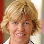 Porträt einer lächelnden Frau mit blonden Haaren, in einem weißen Blazer, vor unscharfem Hintergrund.