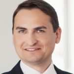 Porträt eines Mannes mit kurzen, braunen Haaren, in Anzug und Krawatte, lächelnd vor hellem Hintergrund.