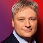 Sönke Rix Porträt eines Mannes mit blonden Haaren, lächelnd vor einem farbigen Hintergrund in Rot- und Violetttönen.