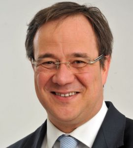 Armin Laschet Porträt eines lächelnden Mannes mit Brille, dunklem Anzug und hellblauer Krawatte vor neutralem Hintergrund.
