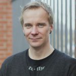 Fabian Wichmann Porträt eines Mannes mit blonden Haaren und Ohrsteckern, der vor einem Zaun steht und in die Kamera lächelt.
