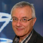 Michael Schroeren Porträt eines Mannes mit Brille, grauen Haaren und einem freundlichen Lächeln, vor einem unscharfen, blauen Hintergrund.