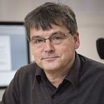 Mann mit Brille und grauen Haaren, trägt einen dunkelgrauen Pullover, sitzt vor einem Computer.
