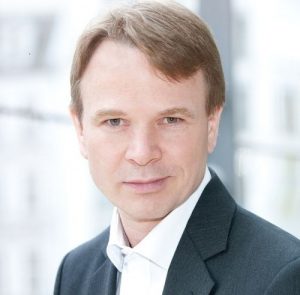 Porträt eines Mannes mit kurzem, braunem Haar, in einem Anzug, vor einem hellen, unscharfen Hintergrund.
