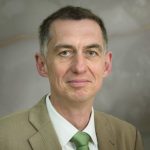 Ralf Beste Porträt eines Mannes mit kurzen Haaren, in einem hellen Anzug und grünem Krawatte, vor einem neutralen Hintergrund.