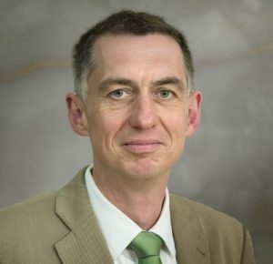 Ralf Beste Porträt eines Mannes mit kurzen Haaren, in einem hellen Anzug und grünem Krawatte, vor einem neutralen Hintergrund.