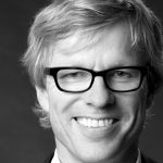 Bernhard Rössler Schwarz-weiß Porträt eines lächelnden Mannes mit Brille und blonden Haaren, vor einem neutralen Hintergrund.