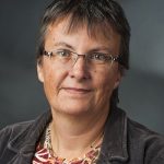 Kathrin Vogler Porträt einer Frau mit kurzen, grauen Haaren, Brille und einem dunklen Blazer, vor einem neutralen Hintergrund.